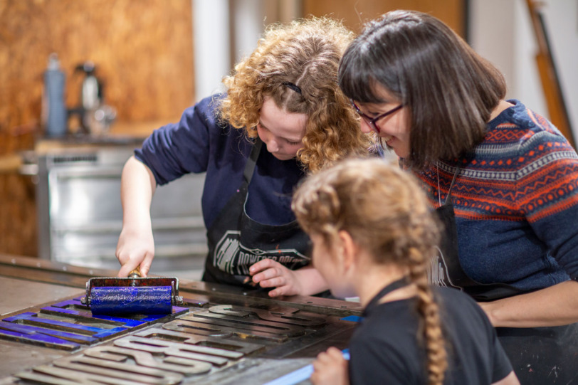 Two children and their mother huddle at the end of the People Powered Press, rolling bright blue ink onto the steel letters